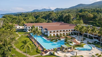 Relax by the expansive pool surrounded by lush greenery and palm trees, with breathtaking views of the ocean from the resort's modern buildings.