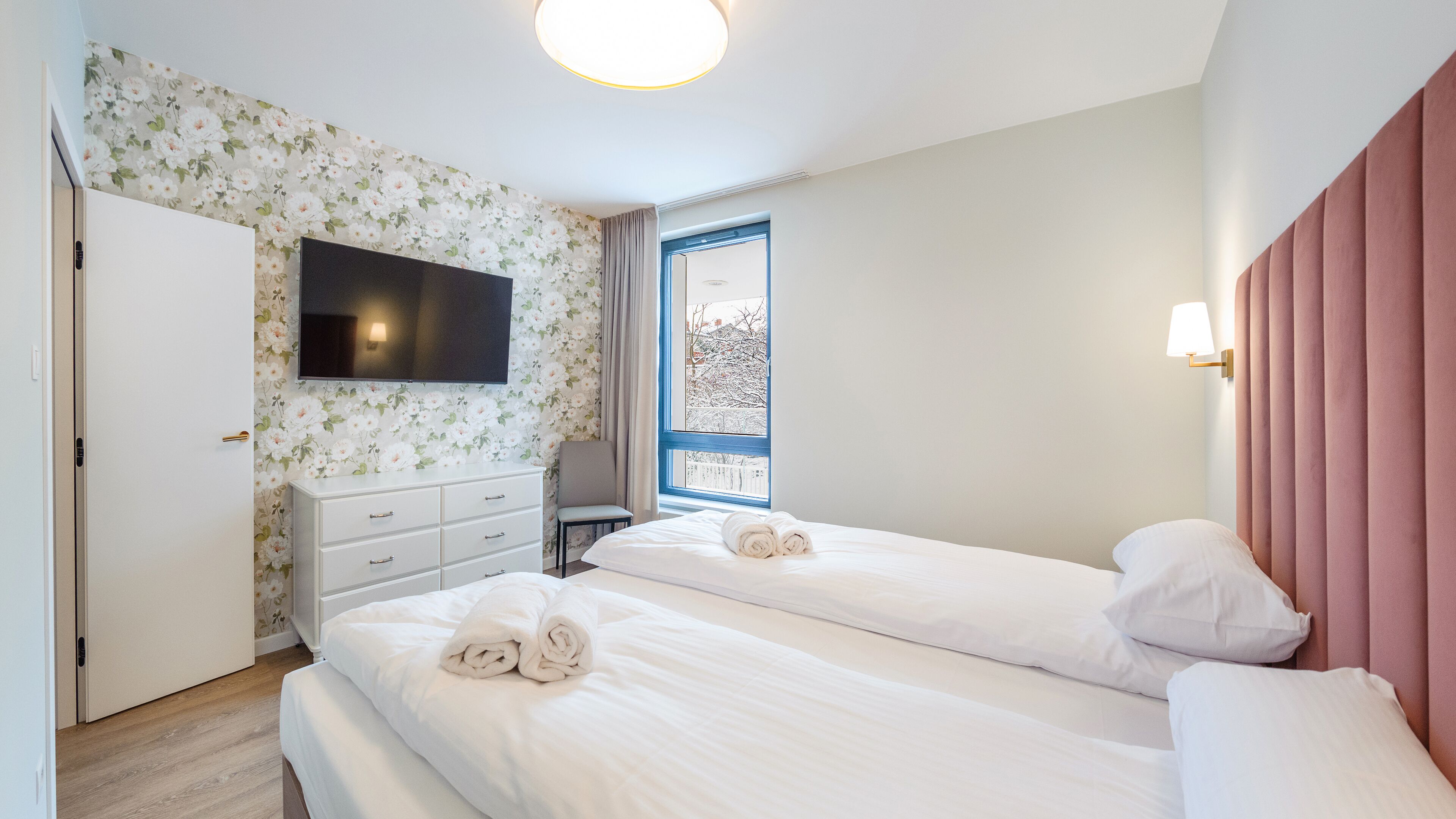 A hotel room with two beds, a TV, a wardrobe, and a window with a view of trees.
