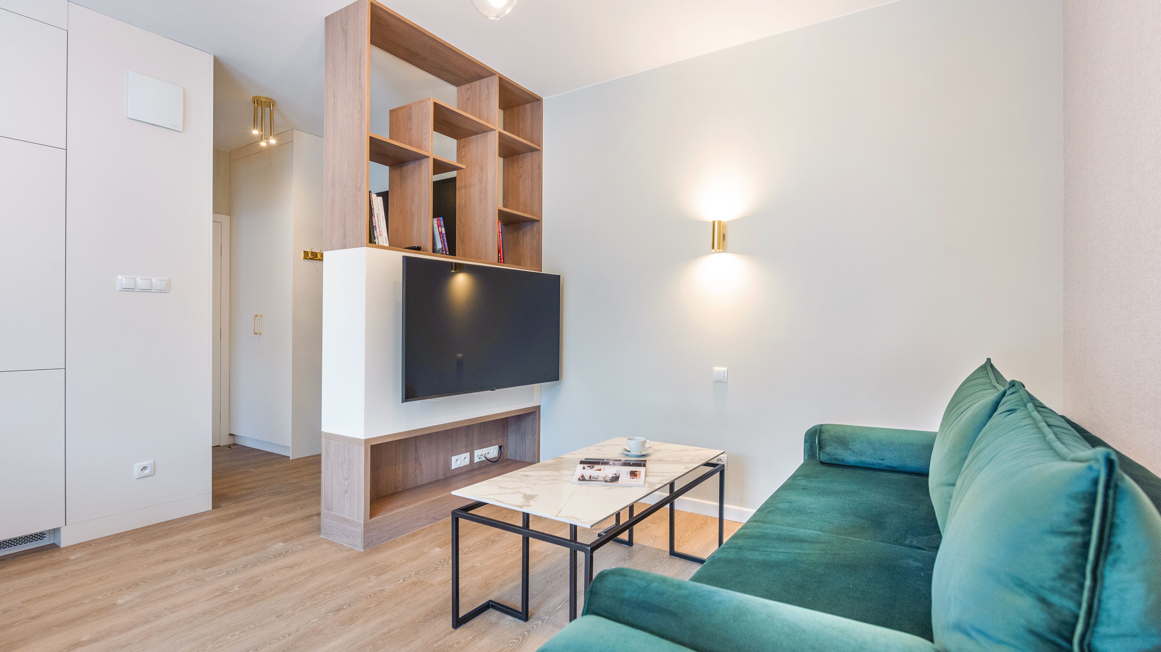 A modern living room with a flat-screen TV, a wooden bookshelf, a green sofa, and a small table with a magazine.