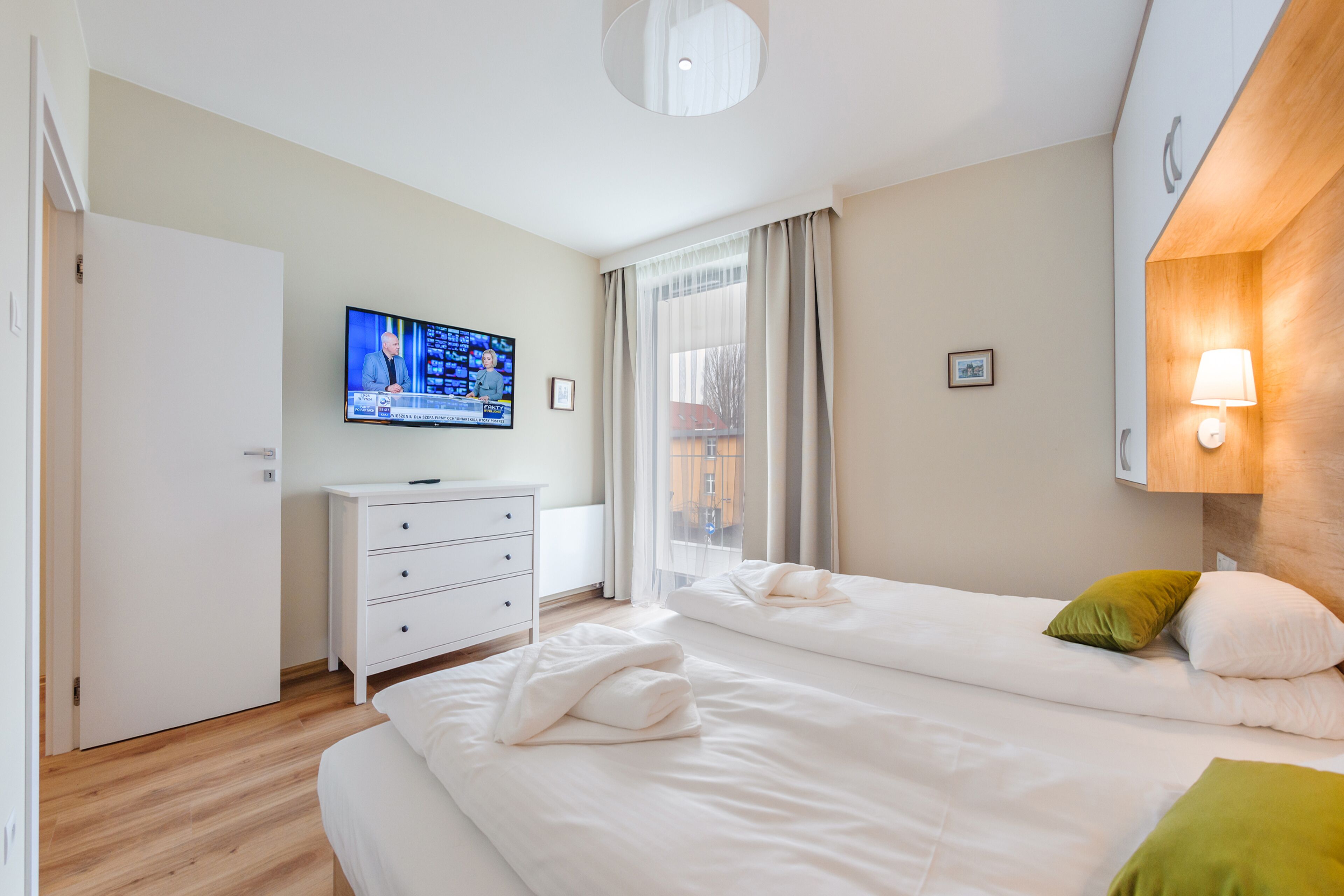 Quarto de hotel com duas camas, televisão na parede, cabeceira de madeira e janela com cortinas.