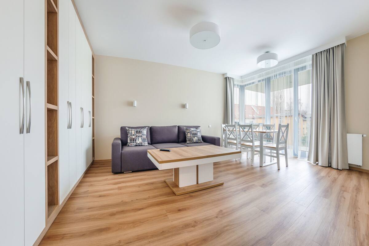 Ein modernes Wohnzimmer mit einer Couch, Couchtisch und Esstisch. Ein großes Fenster mit Vorhängen bietet einen Blick ins Freie.