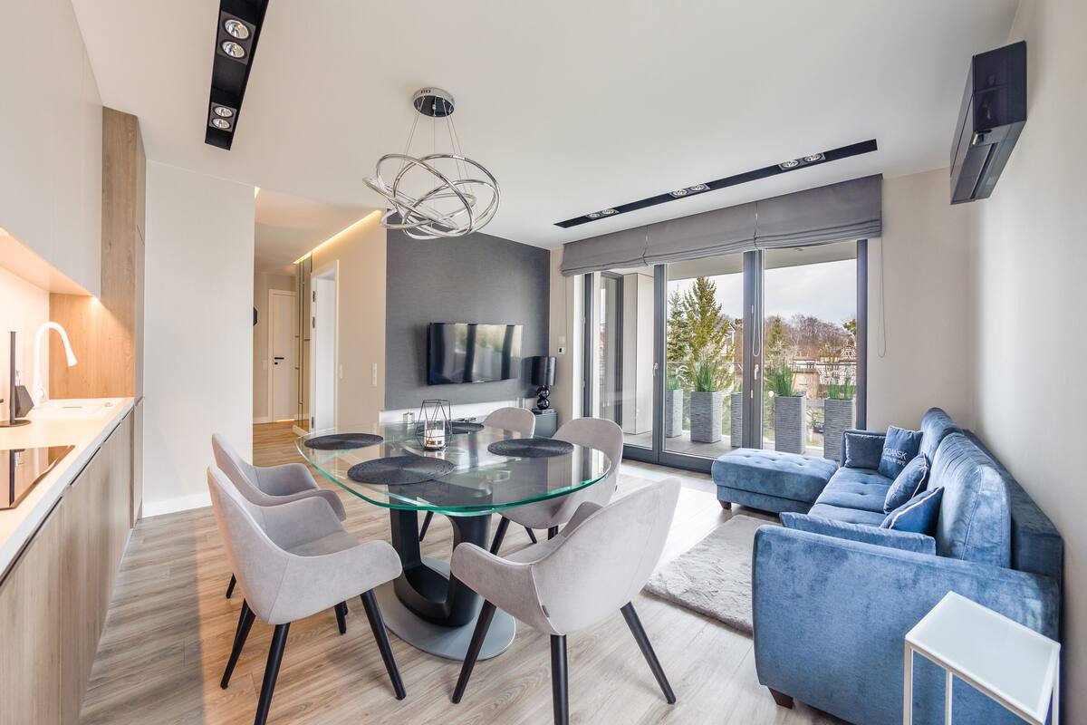 Ein modernes Wohnzimmer mit einem Glastisch, grauen Stühlen, einem blauen Sofa und einem großen Fernseher an der Wand.