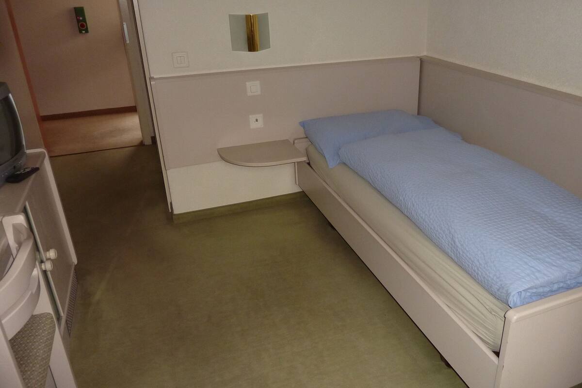 Ein kleines Zimmer mit einem Einzelbett, einem schlichten weißen Bett, einem kleinen Nachttisch und einem Fernseher auf einem Standfuß.