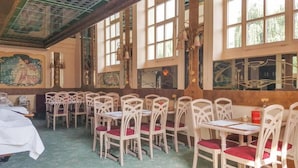 Un espace repas avec des chaises et des tables blanches, de grandes fenêtres et une fresque murale.