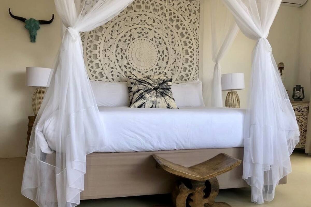 A bedroom with a canopy bed, a decorative headboard, two bedside lamps, and a woven coffee table.