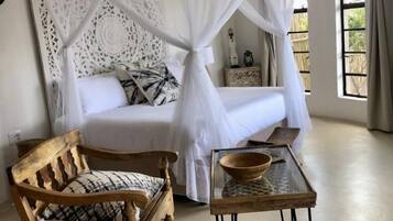 A bedroom with a bed under a white canopy, a wooden chair, a glass table with a bowl, and a patterned rug.