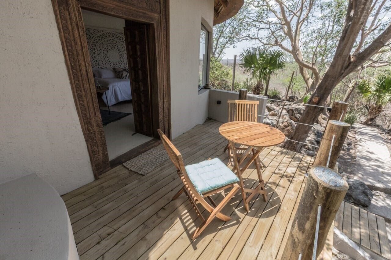 A wooden deck with a table and chairs, a view of a bedroom through an open door, and a natural landscape outside.