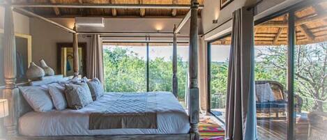 A bedroom with a four-poster bed, a ceiling fan, and a large window offering a view of greenery.