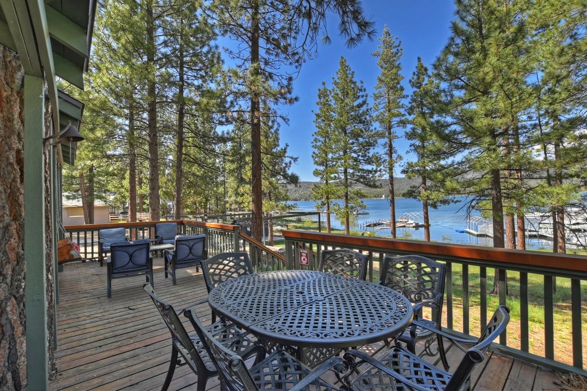 A wooden deck with outdoor seating, overlooking a lake with boats, surrounded by tall trees.