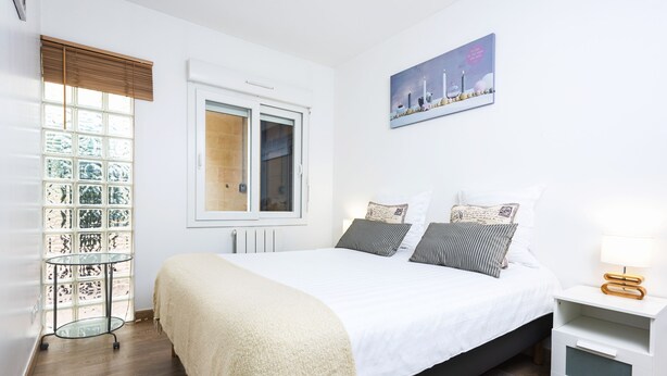 Une chambre à coucher avec un lit, une table de chevet, une lampe, une fenêtre avec des stores et un tableau au mur.