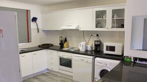A modern kitchen with white cabinets, a dishwasher, a washing machine, a microwave, and a coffee maker. - Bell Rock 19 (Plettenberg Bay)