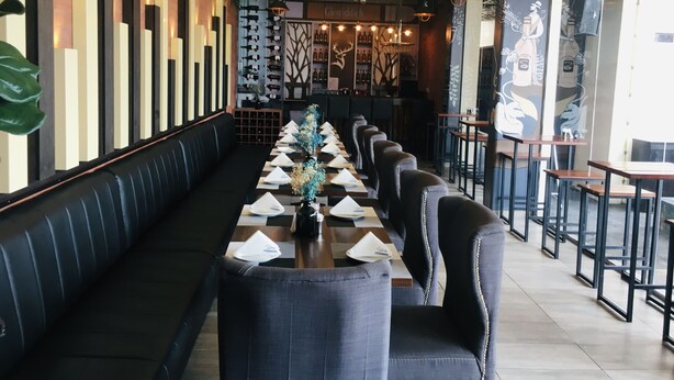 A restaurant with a long table set for dining, black upholstered booths, and a wall with vertical light fixtures and greenery.