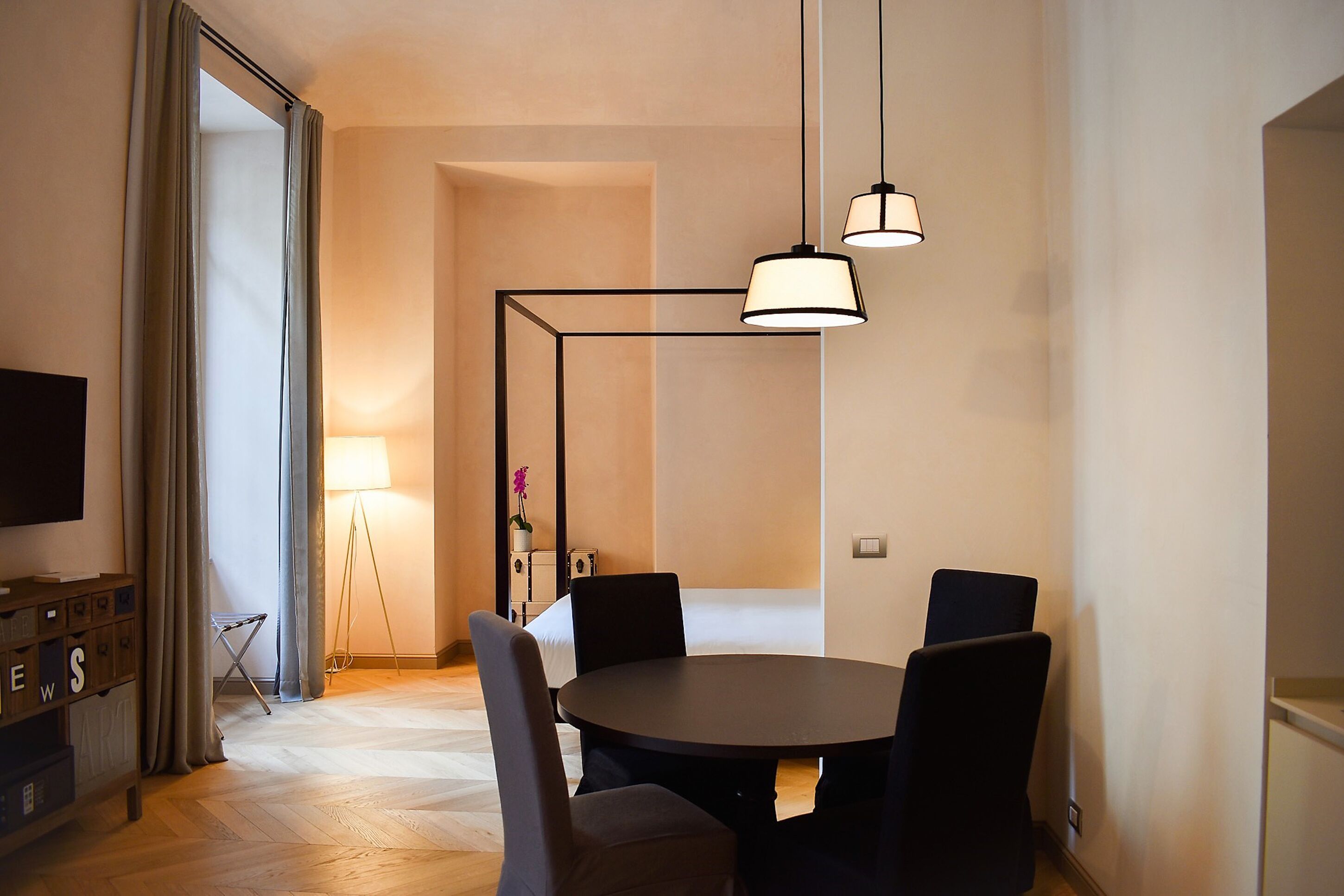 A modern dining area with a round table, four chairs, and pendant lighting. A television is mounted on the wall to the left, and a small table with a lamp and decorative item is visible in the background.