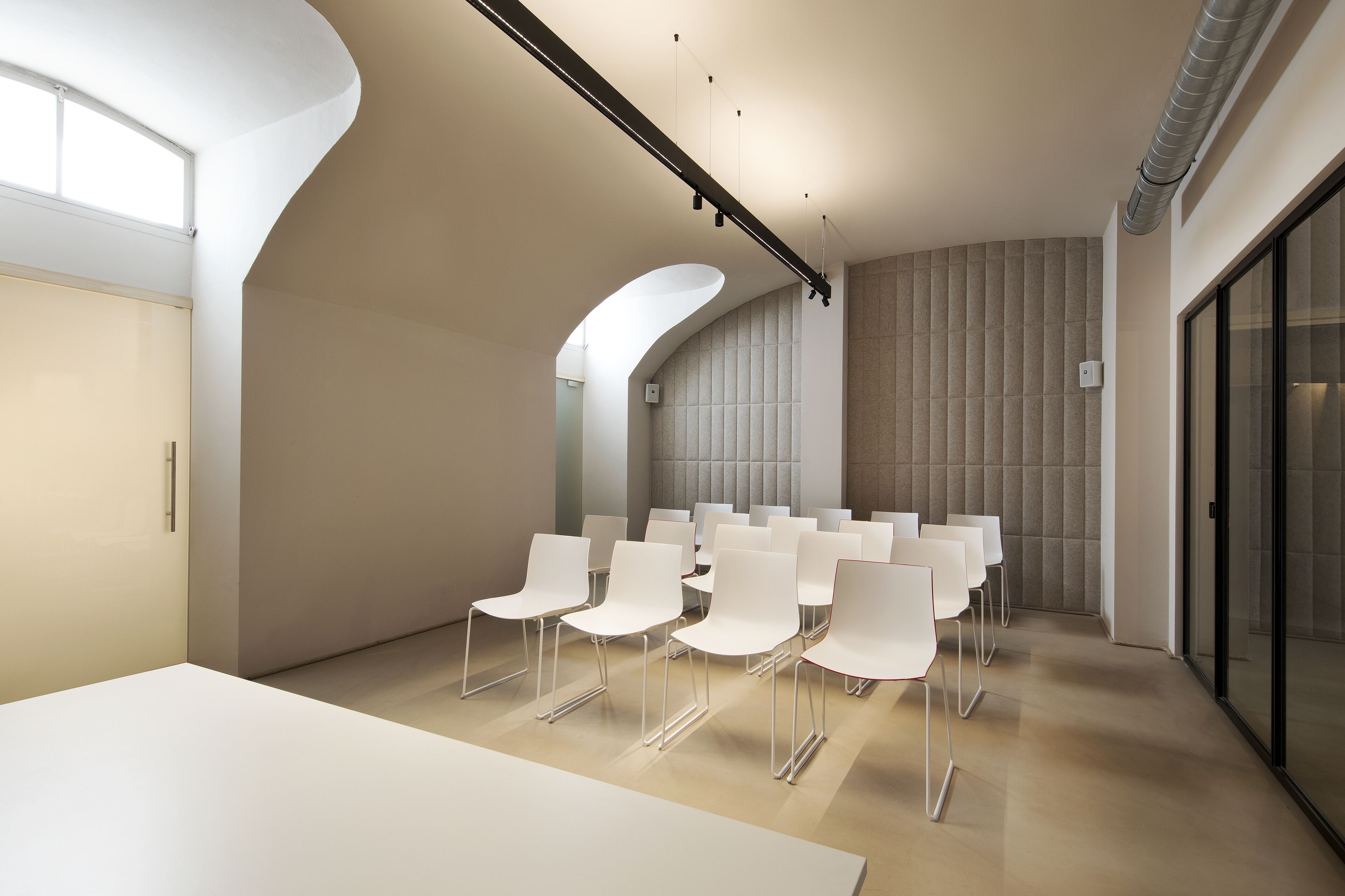 A modern conference room with white chairs arranged in rows, a large white table, and a curved wall with a light-colored panel.