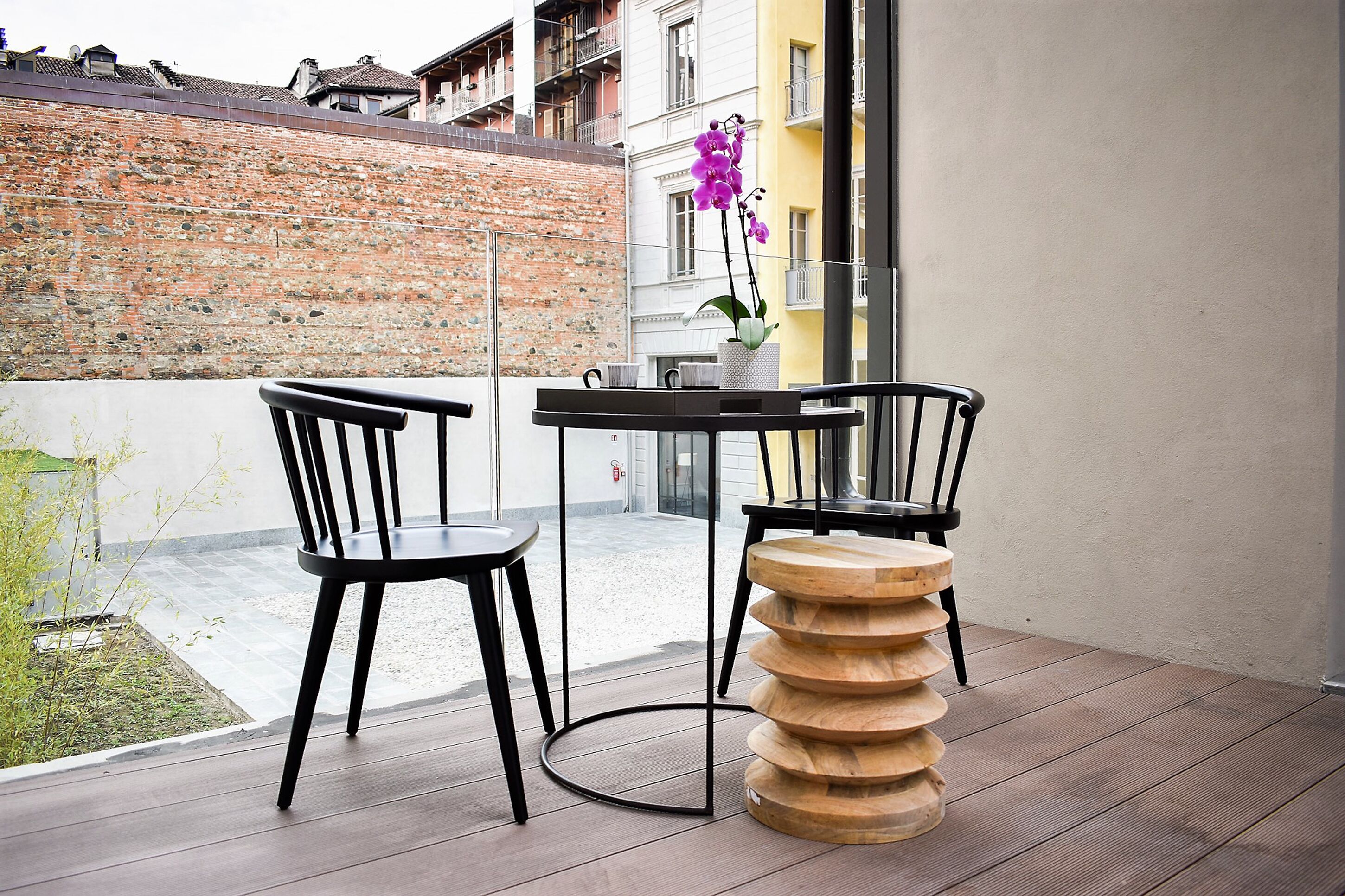 A balcony with two black chairs and a round wooden table, a brick wall, and a building with balconies in the background.