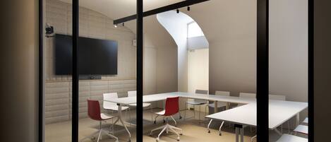 A modern conference room with white tables and red chairs, a flat-screen TV on the wall, and a sloped ceiling.