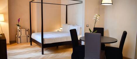 A bedroom with a four-poster bed, a round dining table with chairs, a small table with a vase of flowers, and a hanging light fixture.