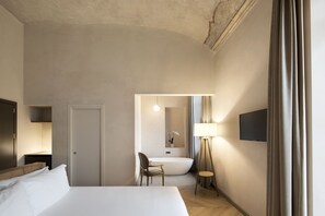 A hotel room with a bed, a chair, a desk, a flat-screen TV, a lamp, and a bathroom with a bathtub and mirror.