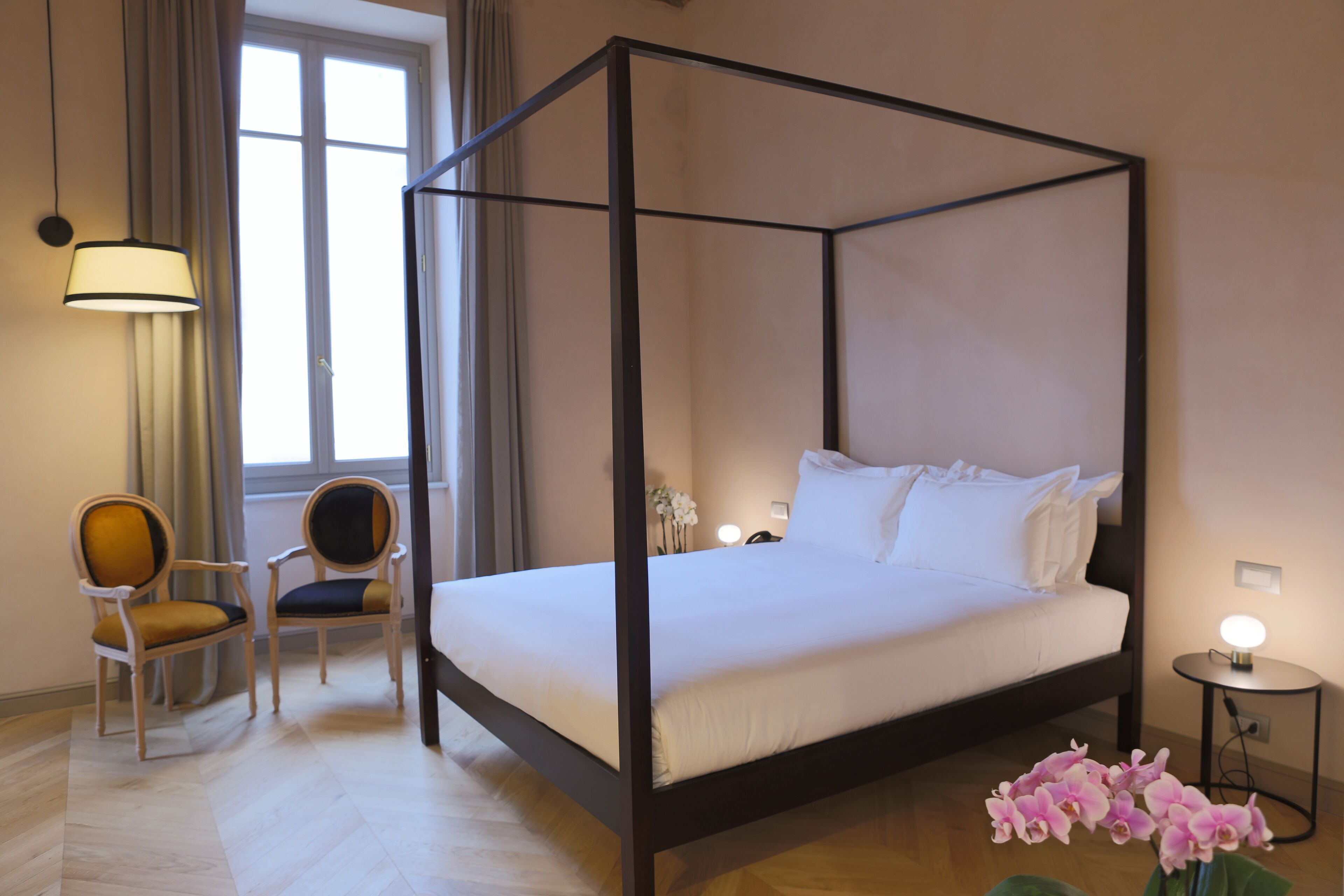 Un letto a baldacchino con lenzuola bianche, due poltrone imbottite, un piccolo tavolino con una lampada e un vaso con fiori rosa.