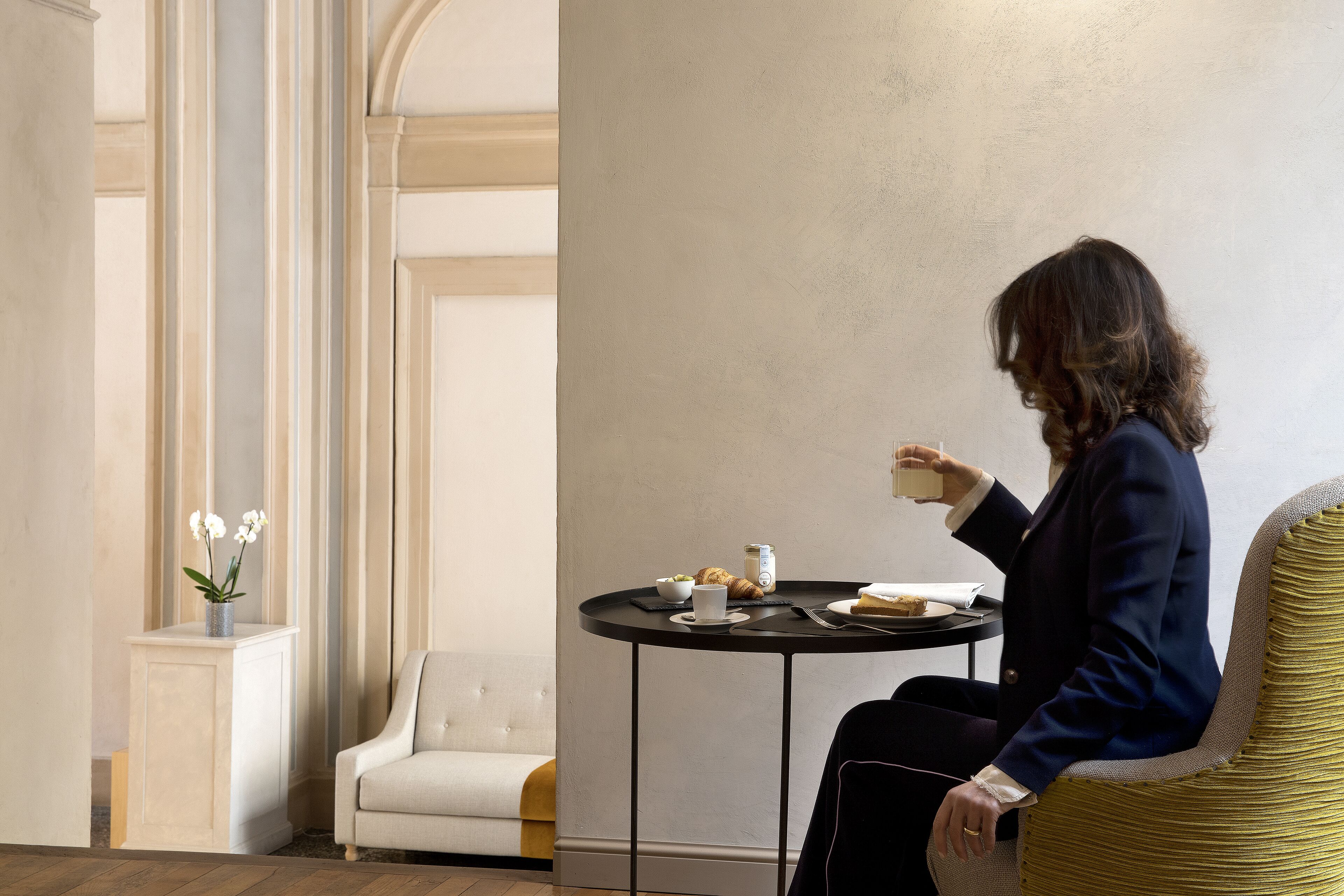 Una mujer sentada a una mesa redonda, con una taza, un plato de comida y un pequeño jarrón con flores.
