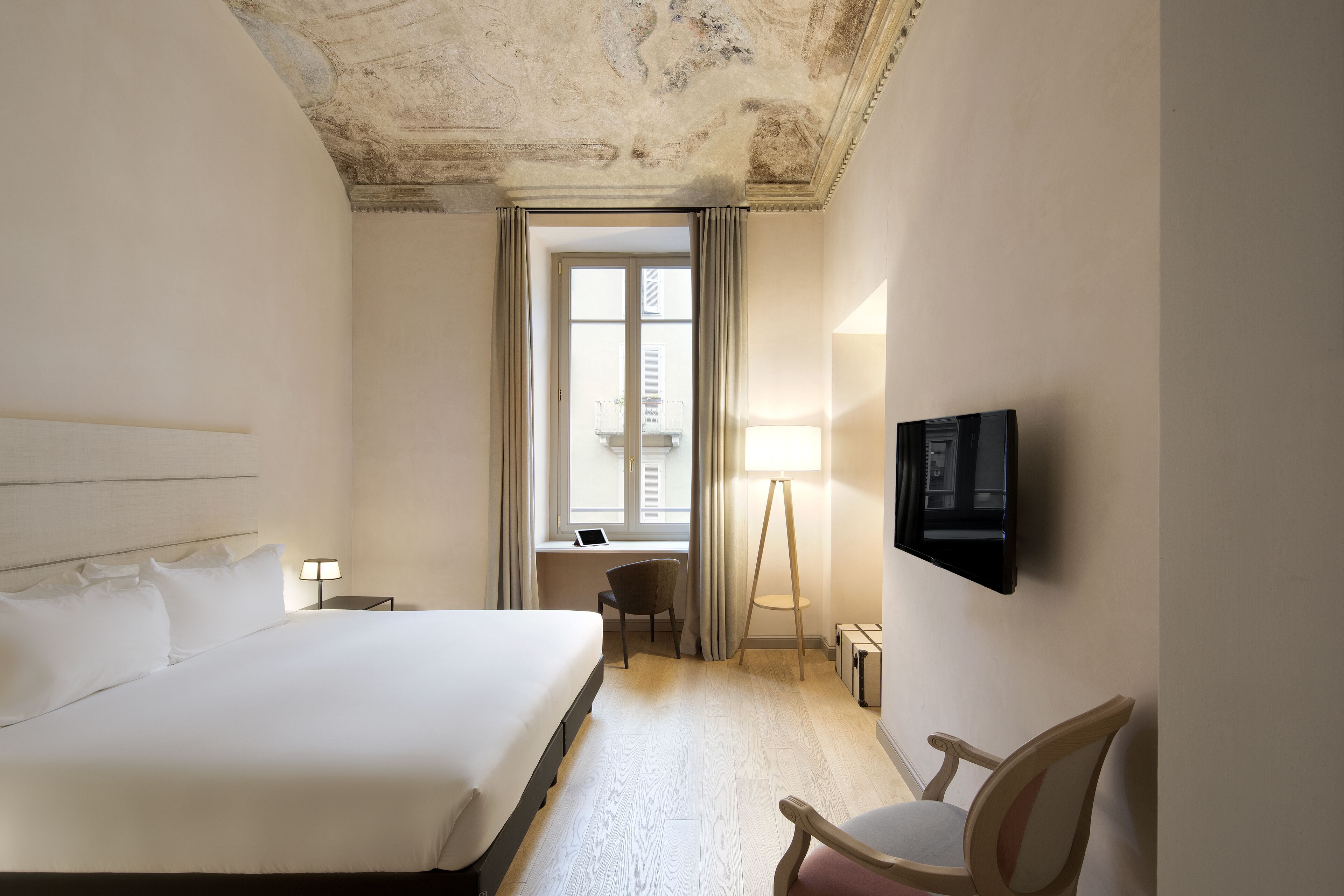Habitación de hotel con una cama grande, un escritorio con silla, un televisor en la pared y una ventana con cortinas.