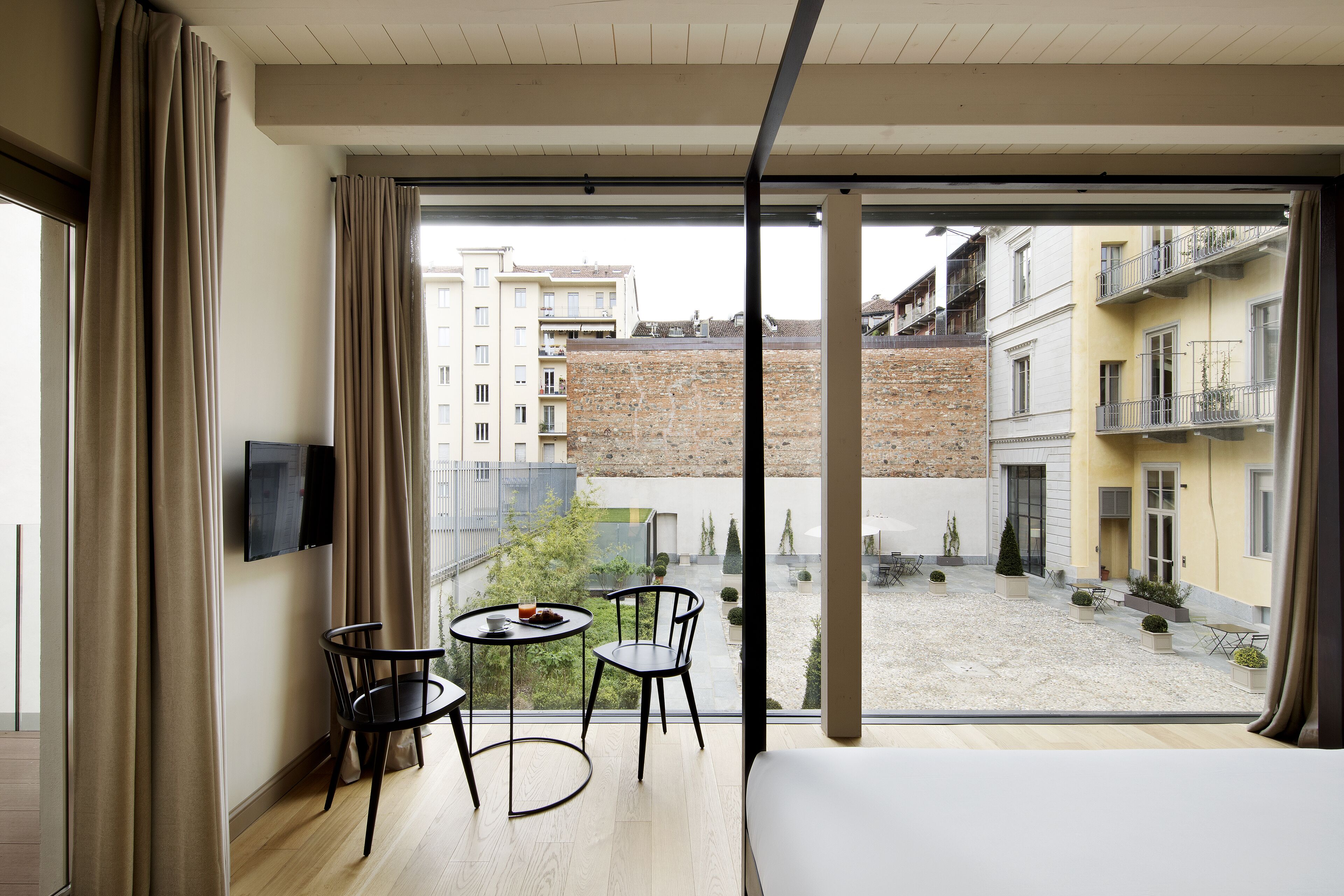 A modern hotel room with a large window, a bed, a TV, and a small dining area with chairs and a table.