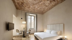 A hotel room with a brick ceiling, a bed with white linens, a round table with chairs, and a TV mounted on the wall.