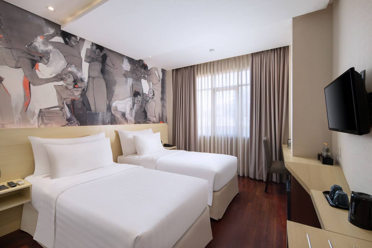 A hotel room with two beds, a large abstract painting on the wall, a TV mounted on the wall, and a desk with a coffee maker.