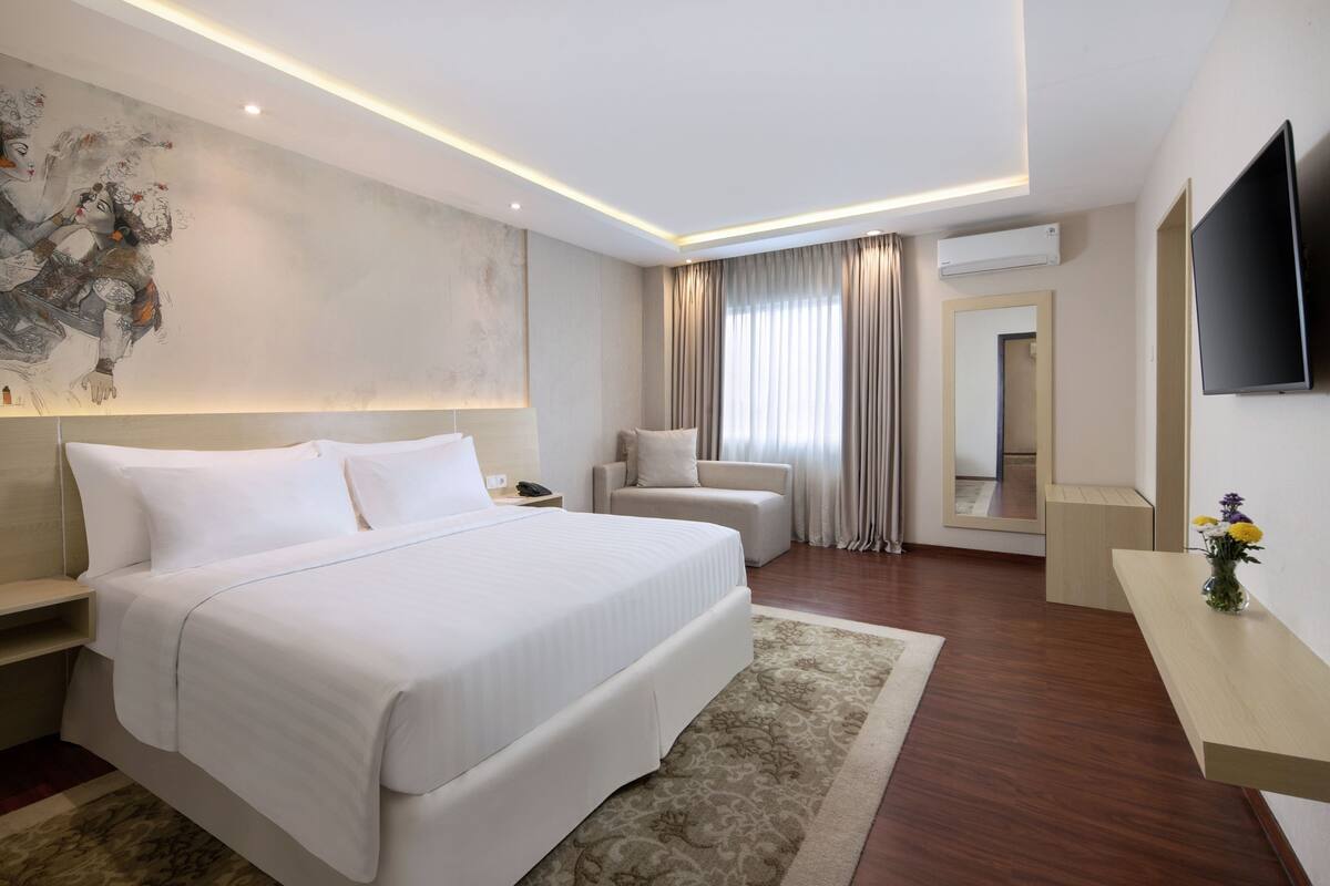 A modern hotel room with a large bed, a sitting area, a flat-screen TV, and a painting on the wall.