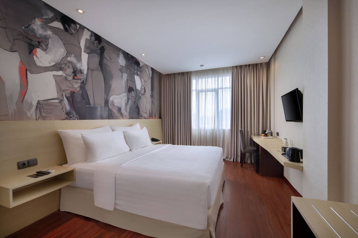 A modern hotel room with a large bed, a flat-screen TV, a desk with a chair, and a large window with curtains.
