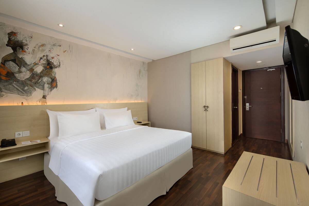 A hotel room with a large bed, a wooden headboard, a flat-screen TV, a wardrobe, and a painting on the wall.
