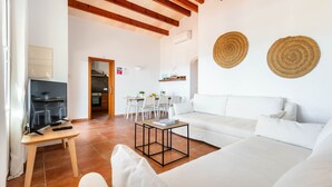 A modern living room with a white sofa, a wooden coffee table, and a TV on a stand. There is a dining area with a table and chairs in the background.