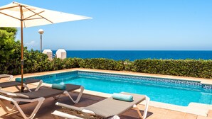 A poolside area with lounge chairs and an umbrella, overlooking a clear blue ocean.