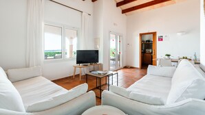 A modern living room with white sofas, a TV on a stand, and a dining area in the background.