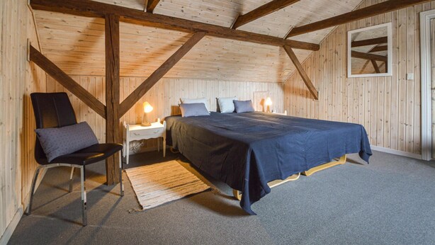 Ein Schlafzimmer mit einem großen Bett, einem Sessel, einem Nachttisch und einer Holzbalkendecke.