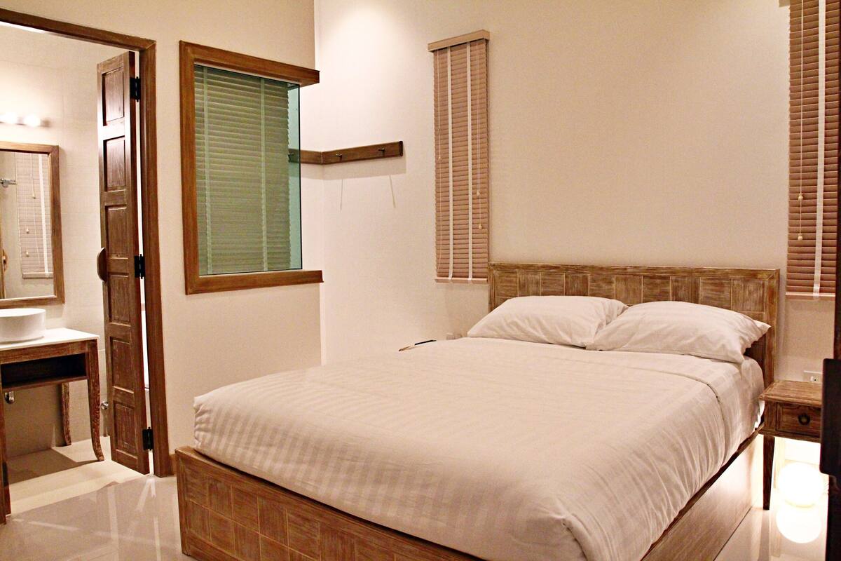 Une chambre à coucher comprenant un lit, une table de chevet, une commode, une tête de lit en bois, une fenêtre avec des stores et une porte donnant accès à une salle de bain.