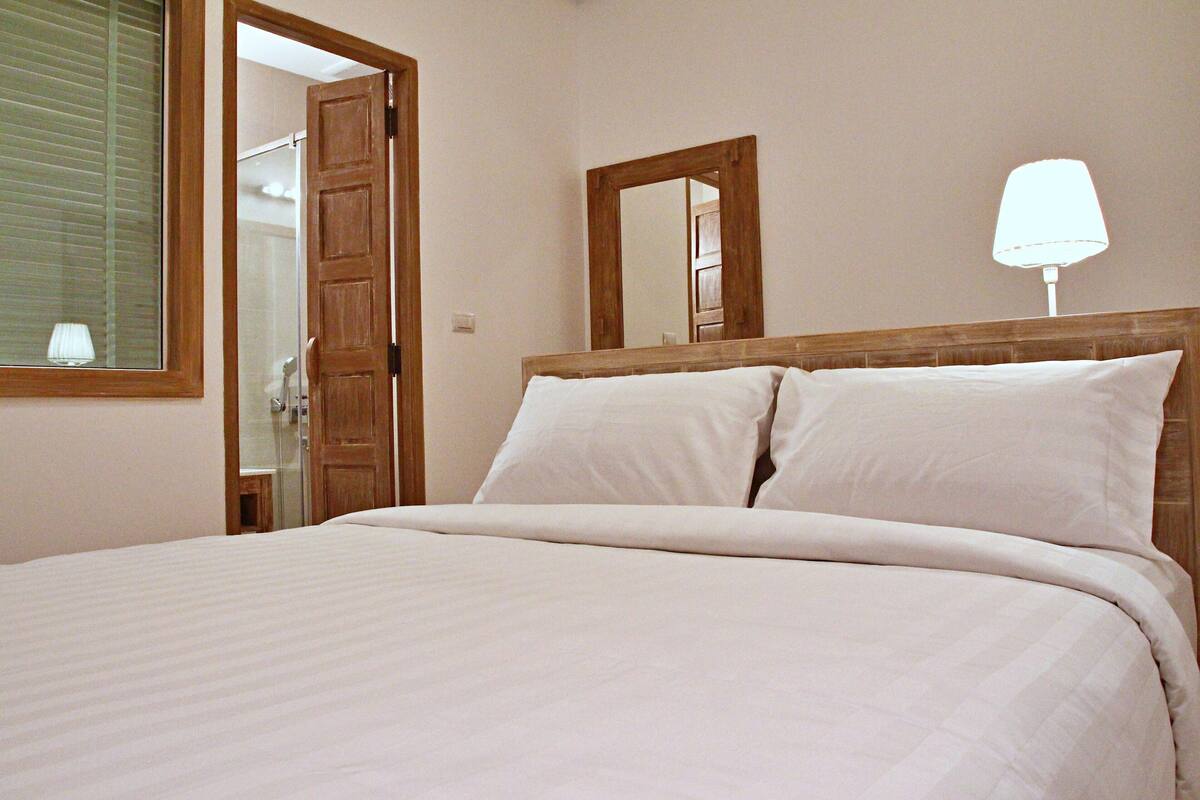 Un lit bien fait, avec du linge de lit blanc, dans une chambre dotée d’une tête de lit en bois et d’un grand miroir au mur.