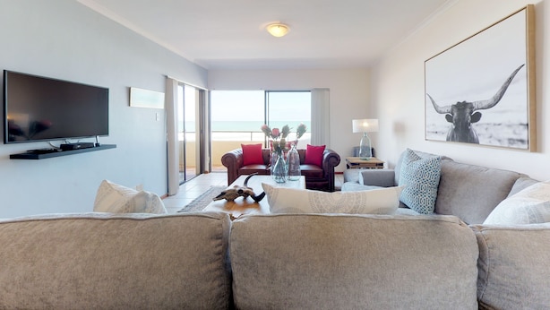 A modern living room with a large flat-screen TV, a grey sofa, a red armchair, and a coffee table with decorative items. - Blouberg Bliss - Beach Condo (Cape Town)