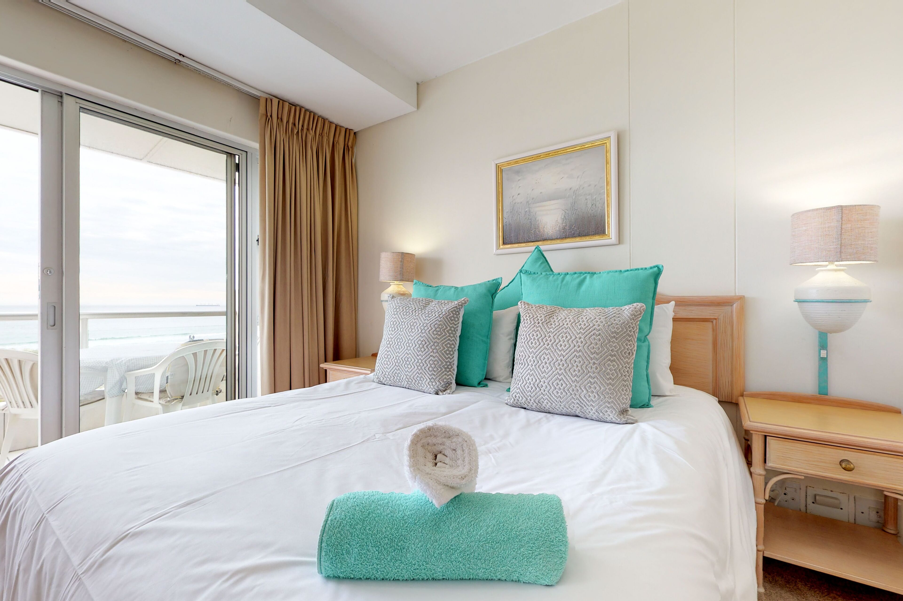 A hotel room with a bed, a view of the sea, a painting on the wall, and a window with curtains.