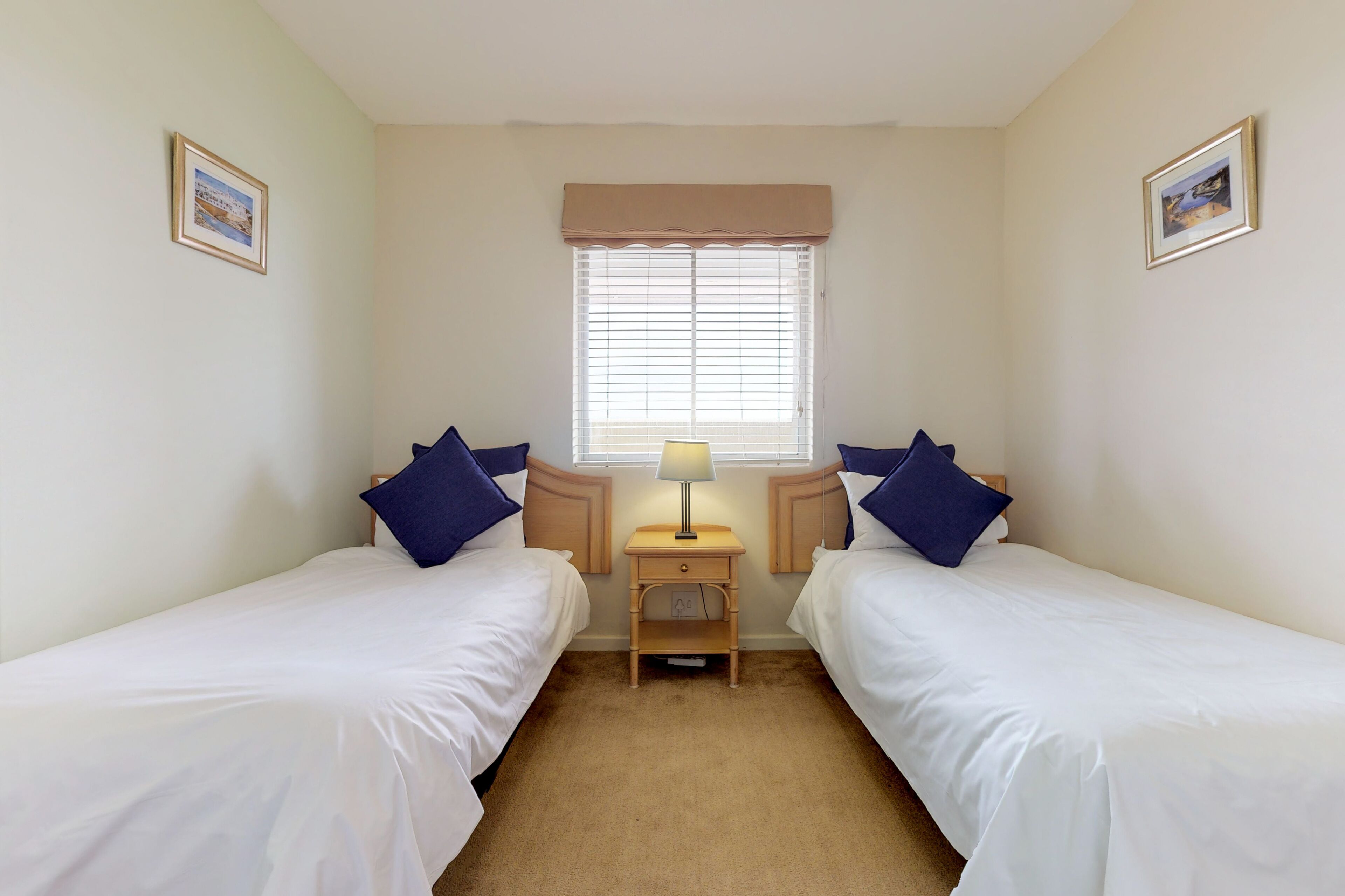 A twin room with two single beds, a bedside table with a lamp, a window with blinds, and two framed pictures on the walls.