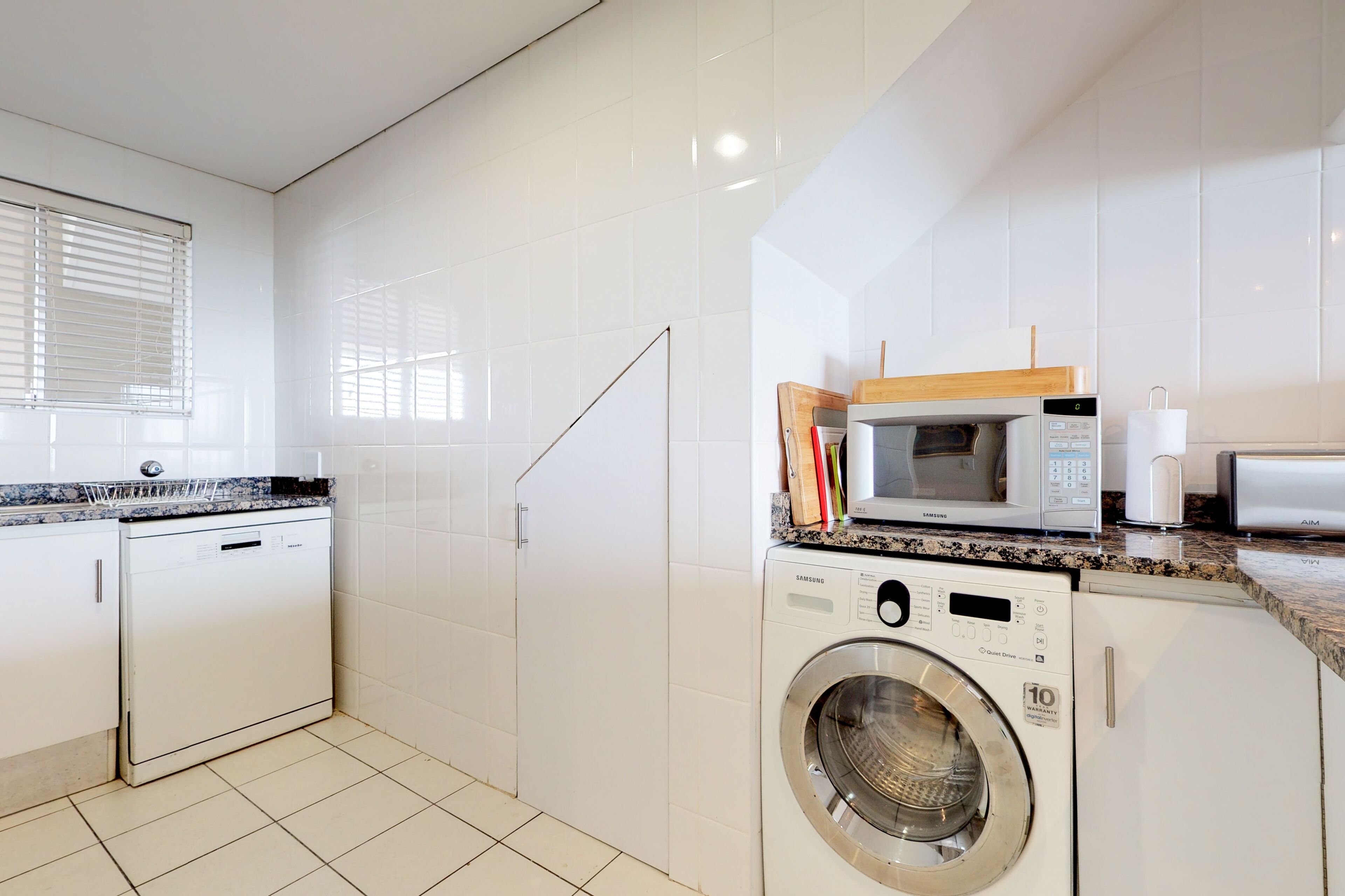 Una cocina moderna con muebles blancos, lavavajillas integrado, lavadora, microondas y tostadora.