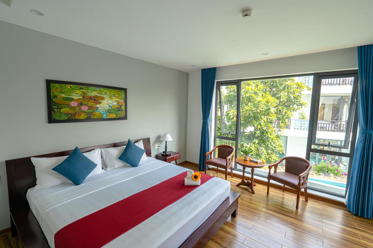 A hotel room with a large bed, a painting on the wall, a balcony with a view of greenery, and a small table with chairs.