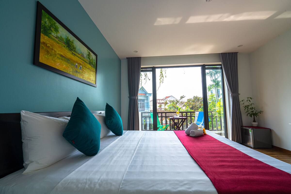 A hotel room with a bed, a painting, a balcony with a view, and a plant.