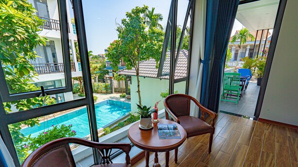 A balcony with a view of a swimming pool, two wooden chairs, a small table with a magazine, and a potted plant. - Golden Star Villa Hoi An (Da Nang)