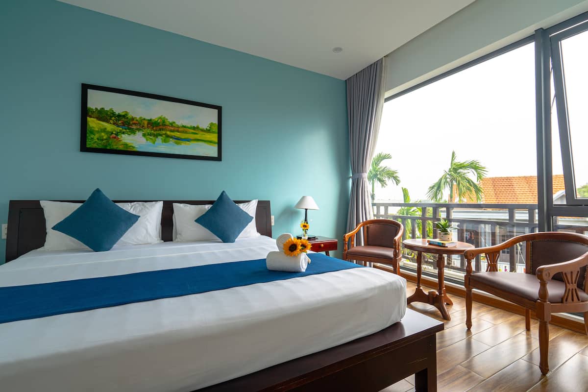 A hotel room with a large bed, blue bedding, a painting of a landscape, a balcony with a view of palm trees, and a wooden chair.