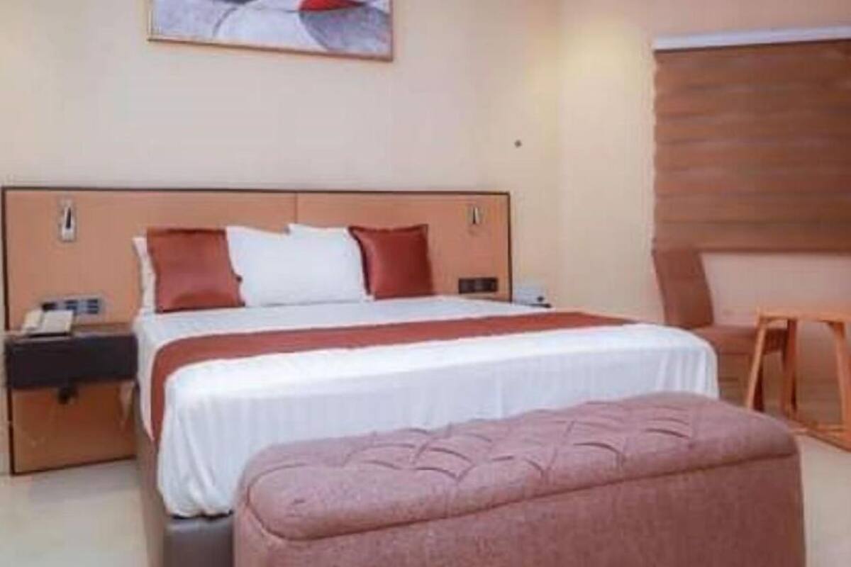 Hotellihuone, jossa on sänky, yöpöytä, tuoli, ikkuna, jossa on sälekaihtimet, ja taulu seinällä.