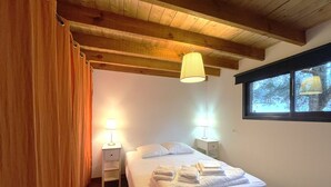 Ein Schlafzimmer mit einem Bett, zwei Nachttischen mit Lampen, Holzbalken an der Decke und einem Fenster mit Blick auf Bäume.