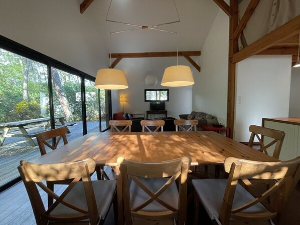 Un vaste espace dédié à la salle à manger, avec une grande table et des chaises en bois, deux lampes suspendues et une vue sur l’extérieur.