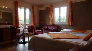 A hotel room with a bed, a desk, a chair, and a sofa. There are windows with curtains and an air conditioning unit on the wall. - Hôtel Charles (Les Arcs-sur-Argens)