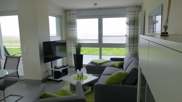 A modern living room with a flat-screen TV, grey sofa with green cushions, a small coffee table, and a potted plant. - Residenz Windjammer (Cuxhaven)